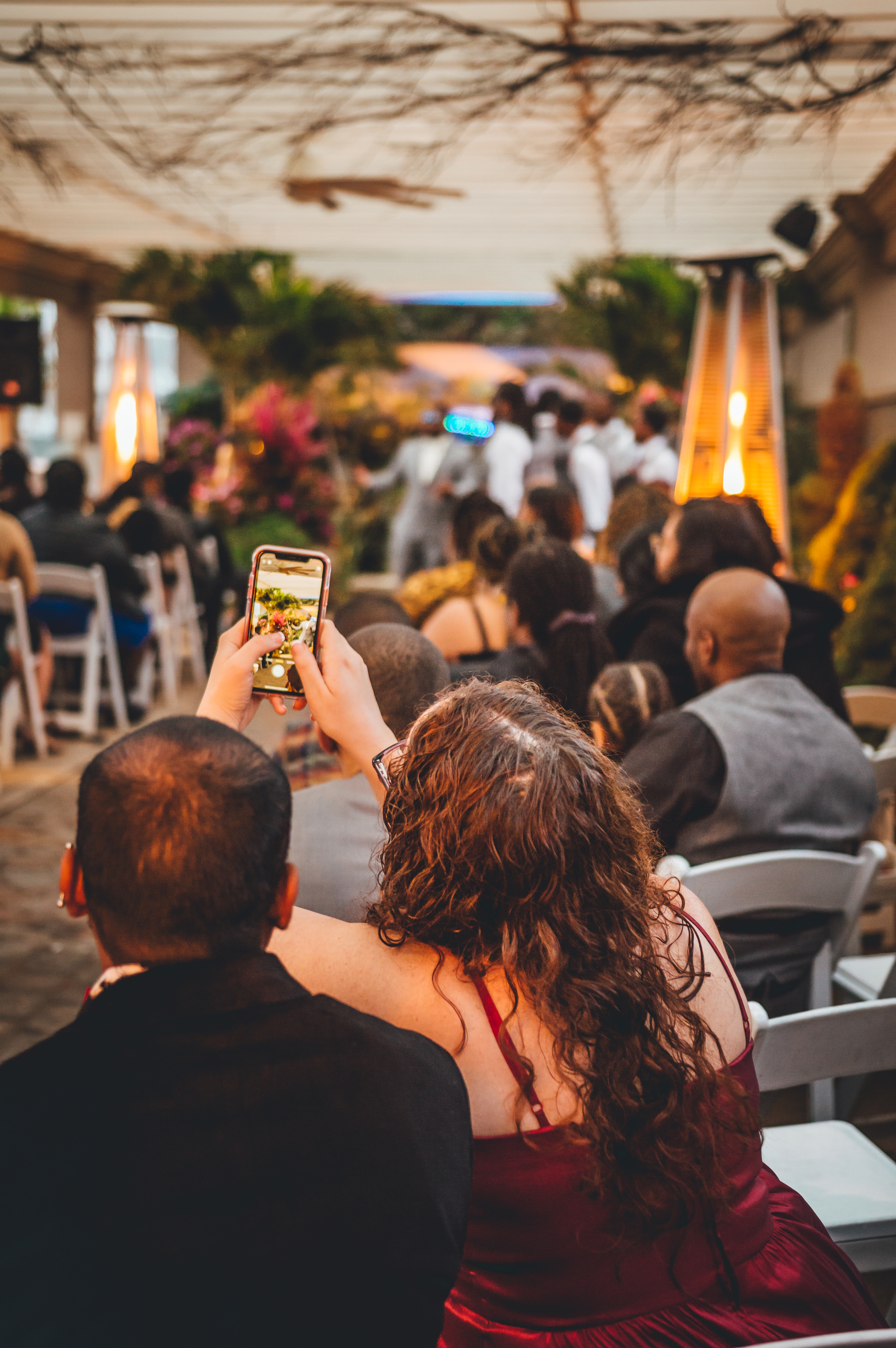 A guest in a burgundy dress photographs the wedding reception with a smartphone, seated among other attendees. The outdoor venue is decorated with amber lantern