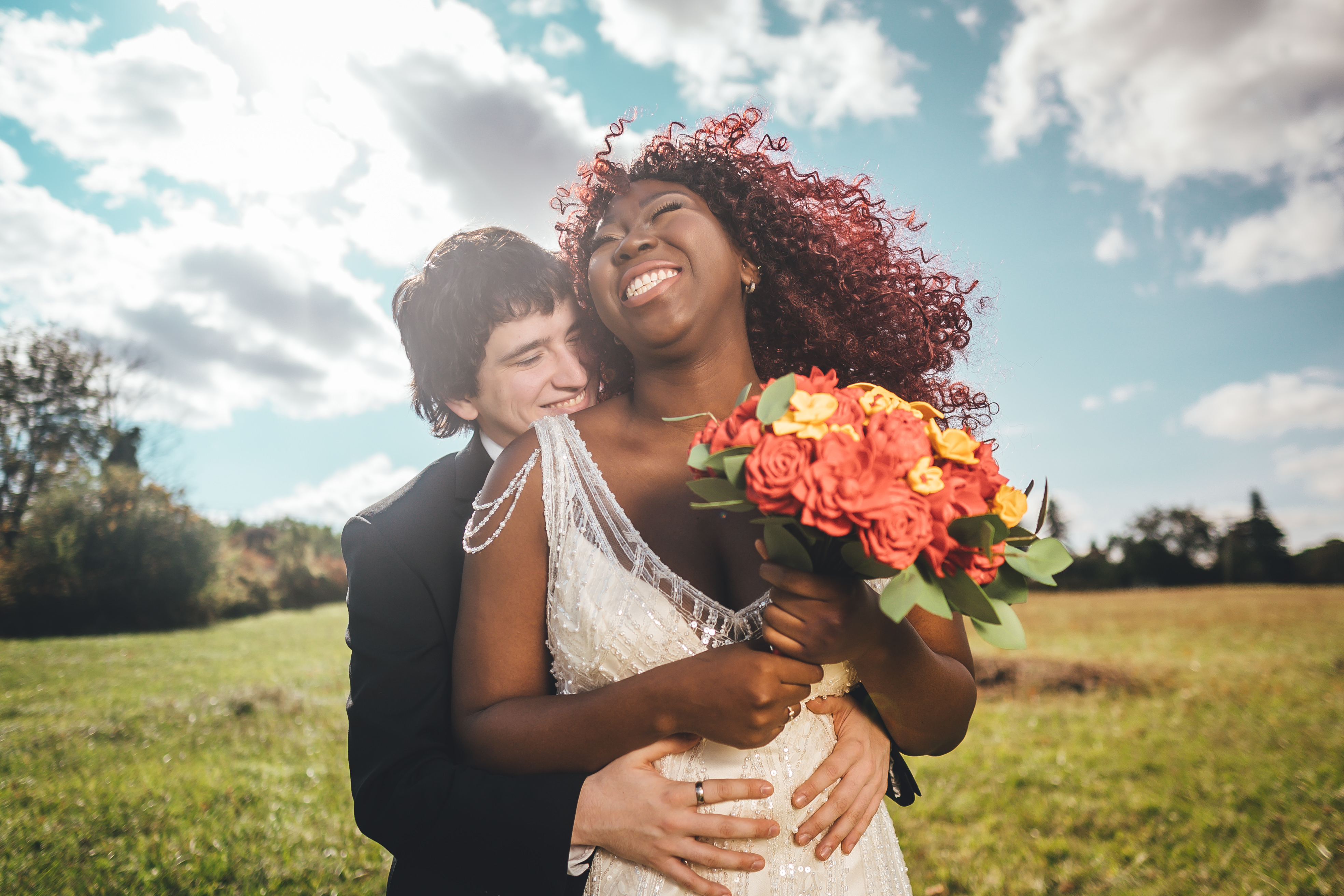 A bride with voluminous curly red hair in a white lace halter dress holds a vibrant coral and yellow bouquet while being