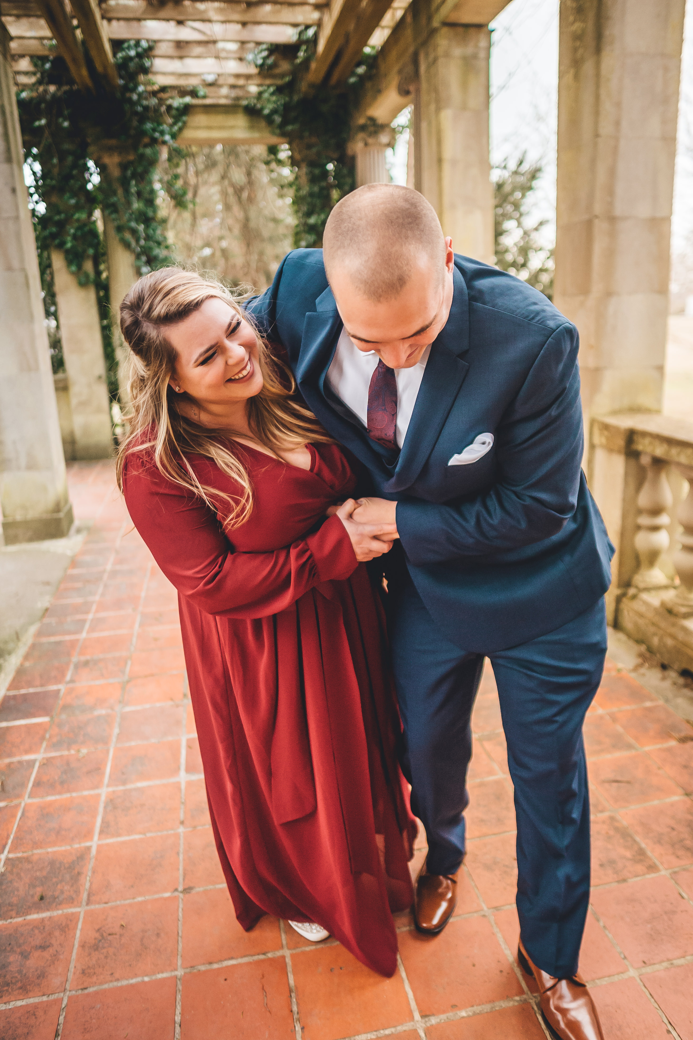 The couple walks together through a classical stone colonnade with ivy-covered pergola overhead, both laughing and smili