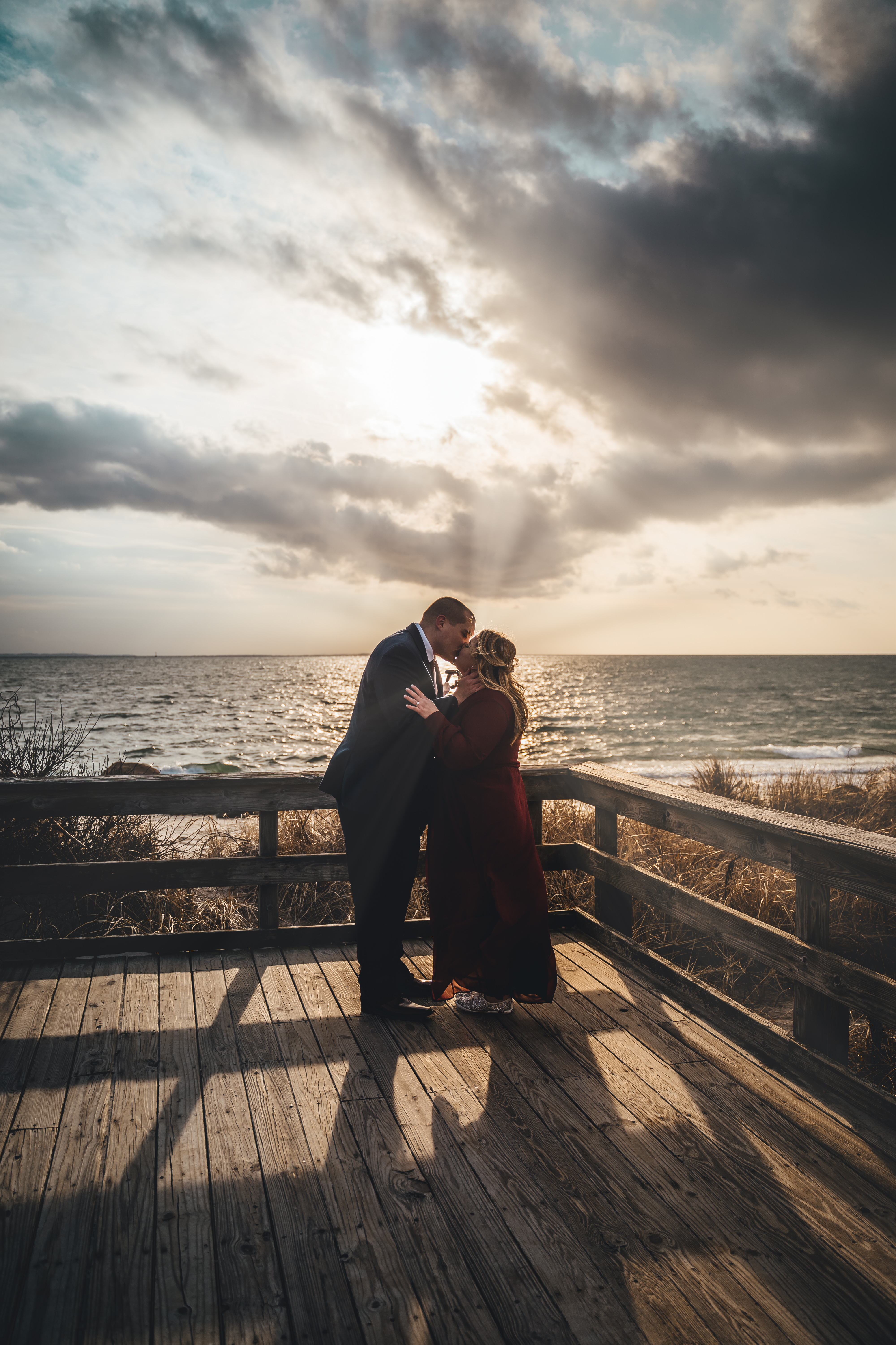 The groom in a dark suit kisses the bride in a burgundy dress on a wooden beach boardwalk, with dramatic storm clouds an
