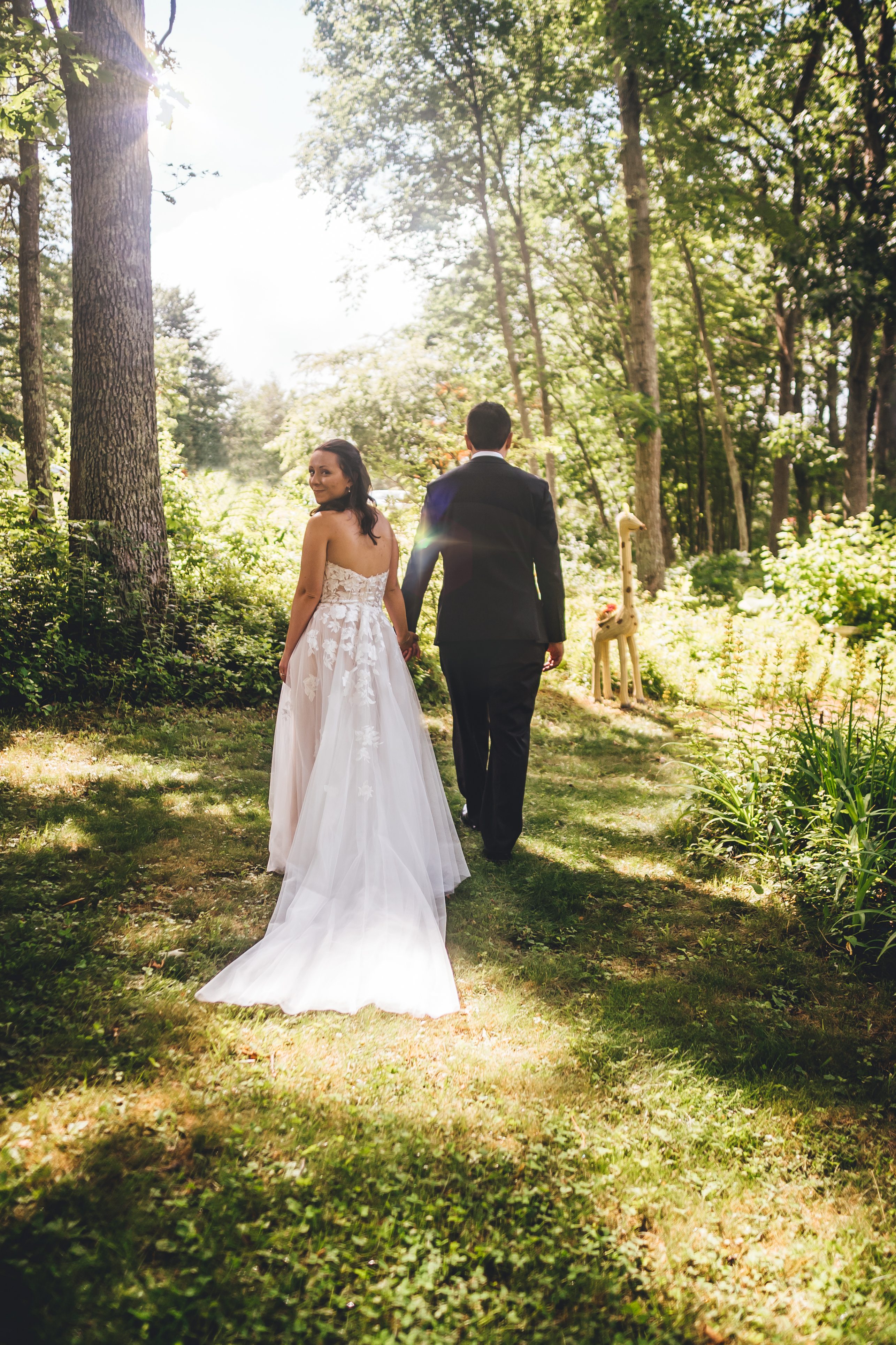 The bride and groom walk together down a tree-lined path in a forest setting, with dappled sunlight filtering through the canopy. The bride wears a flowing whit