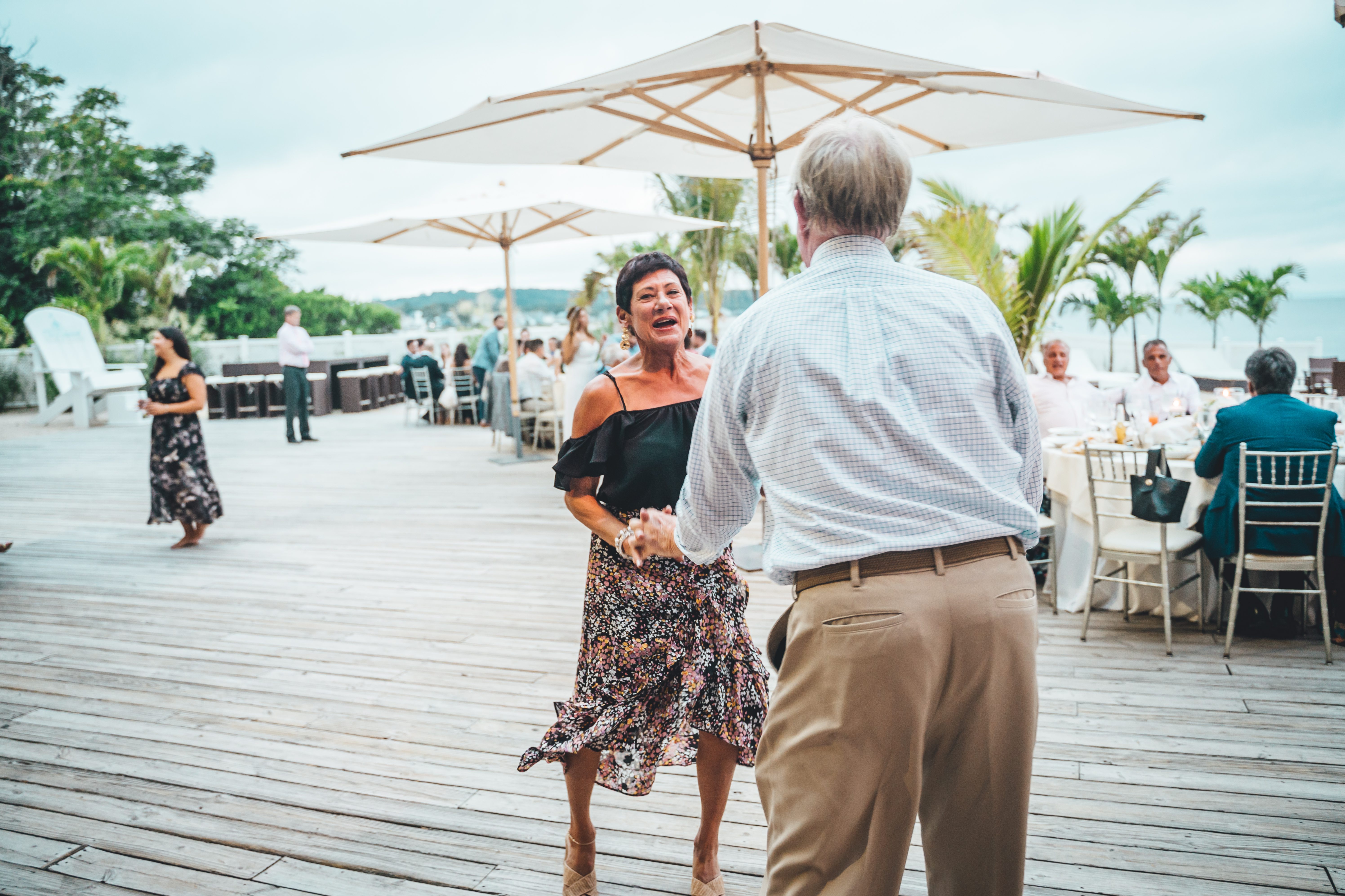 A guest in a black off-shoulder top and patterned skirt dances with an older gentleman in a light blue checkered shirt and tan pants on a wooden deck reception 
