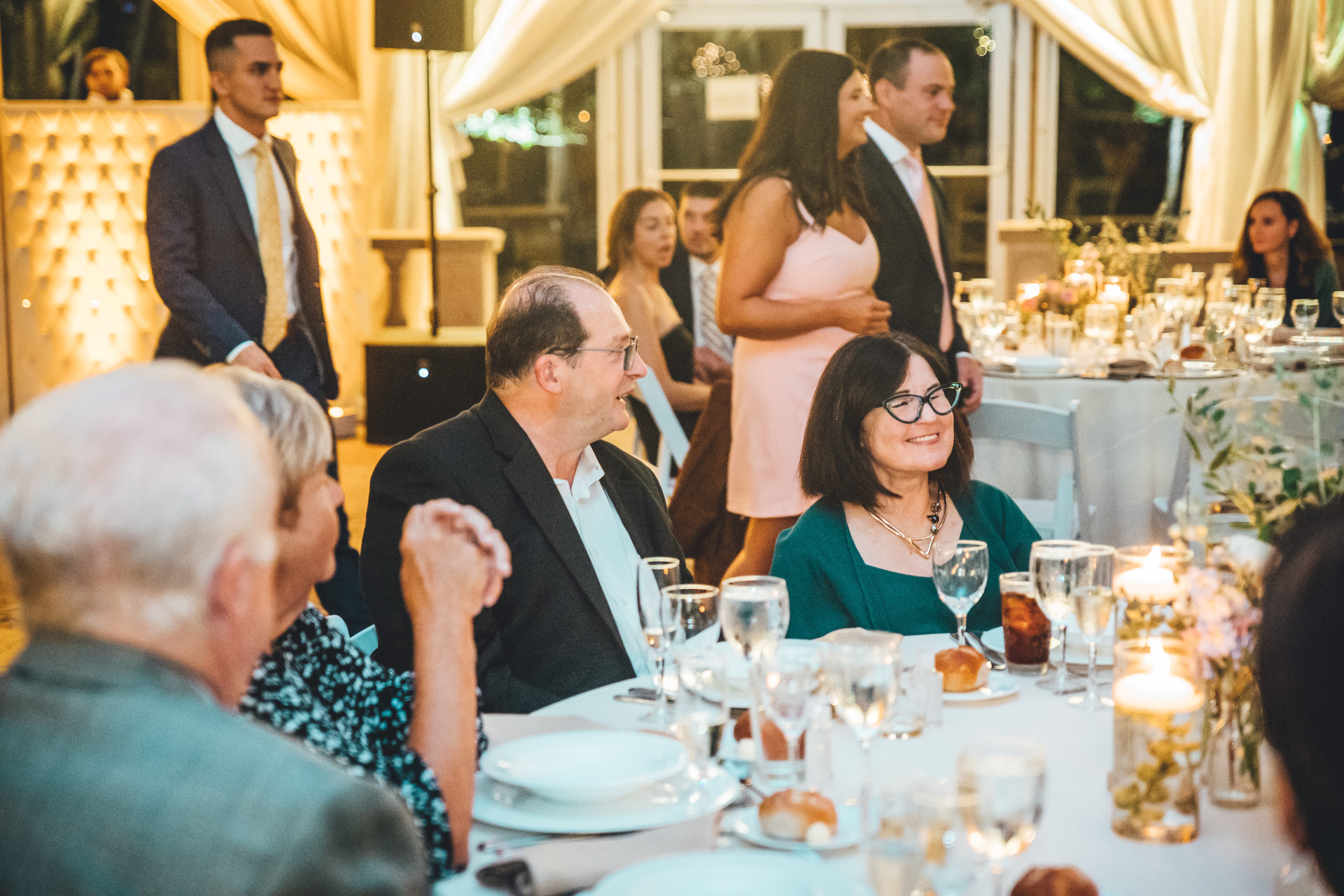 Reception dinner in full swing with guests seated at elegantly dressed tables. The room features warm golden lighting, cream draping, and floral centerpieces. M