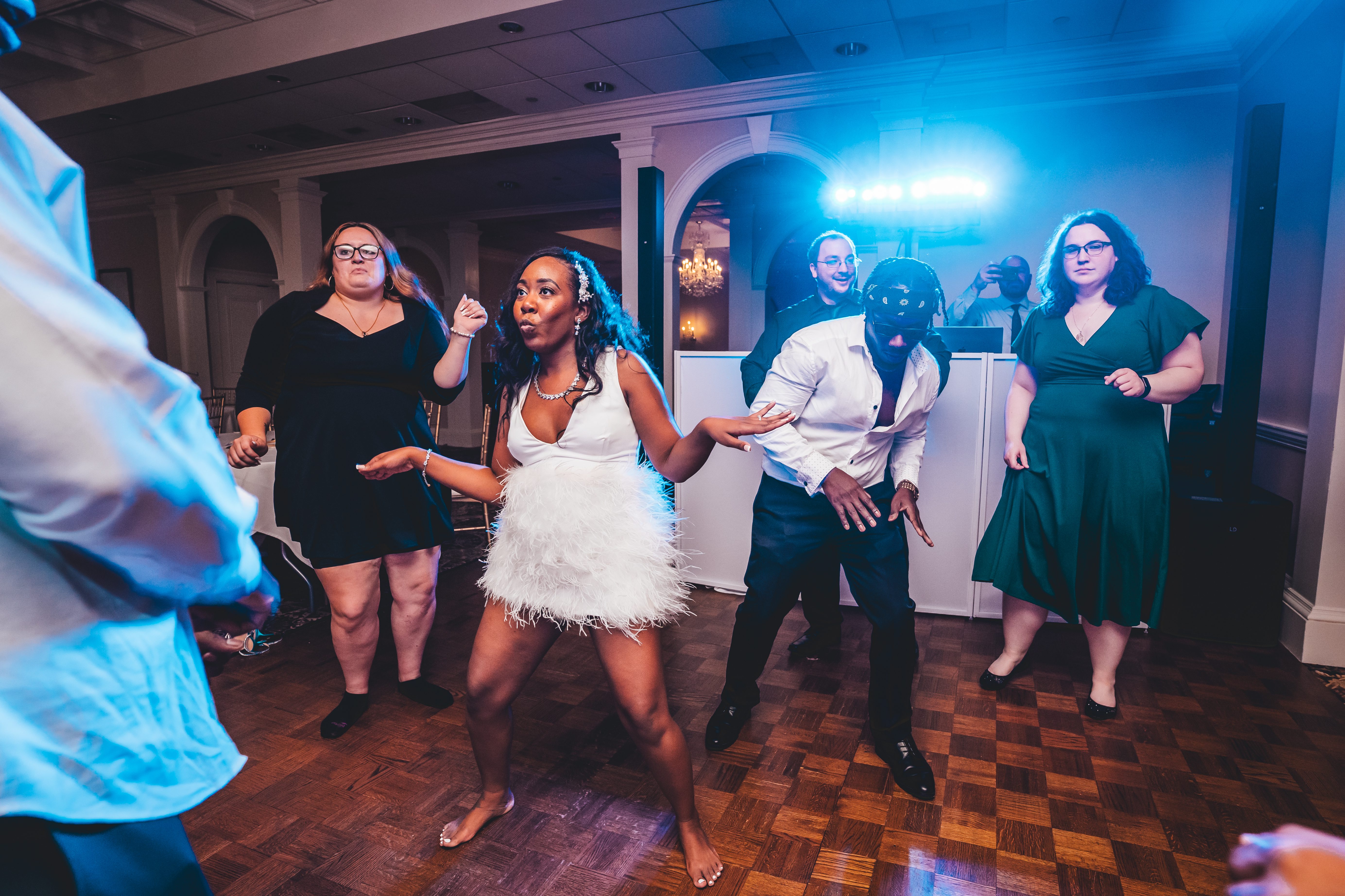 The bride in a white feathered dress dances energetically with wedding guests on a wooden parquet dance floor. A groom f