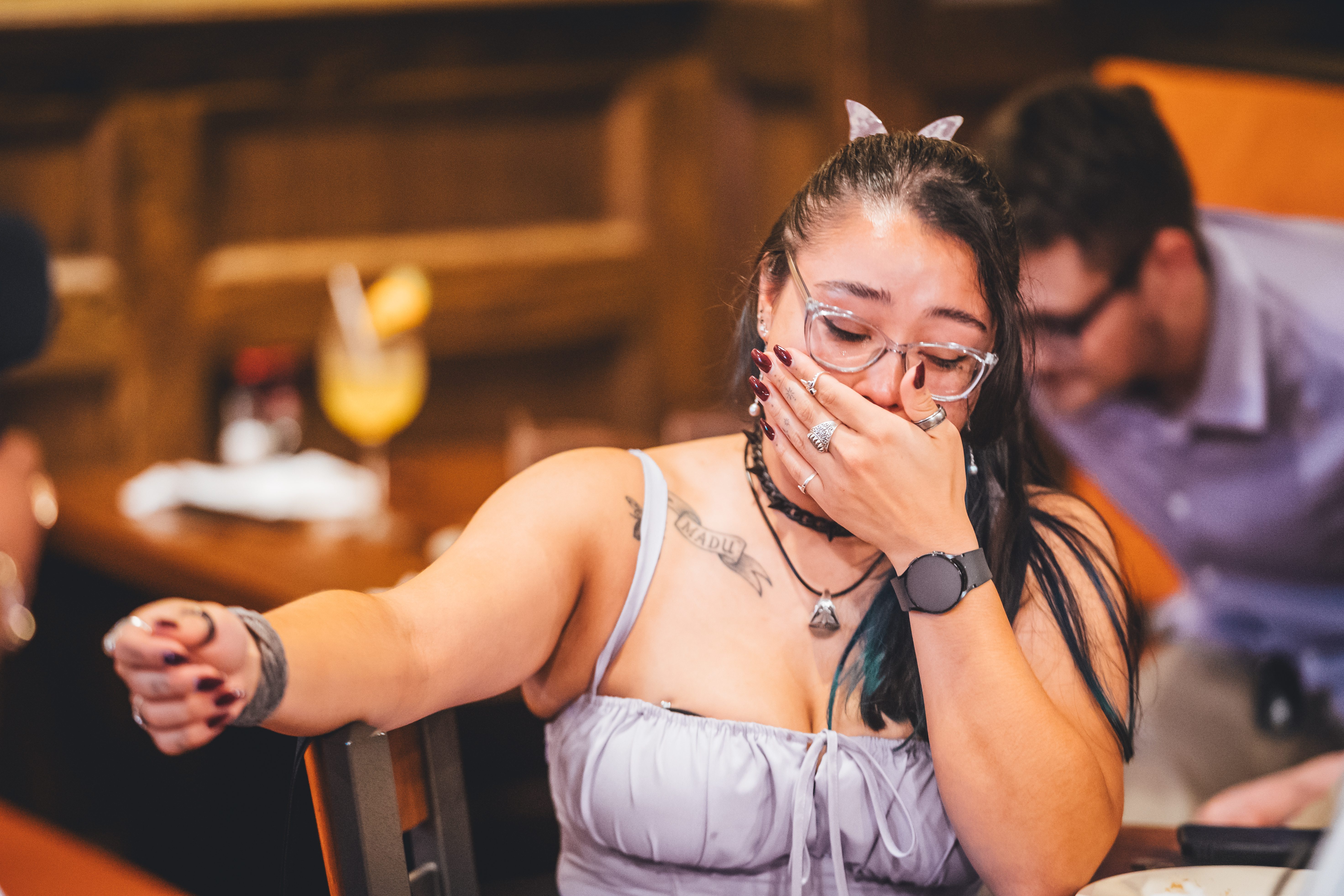 A woman with glasses and white cat-ear headband is laughing with her hand to her face during a reception moment. She wea