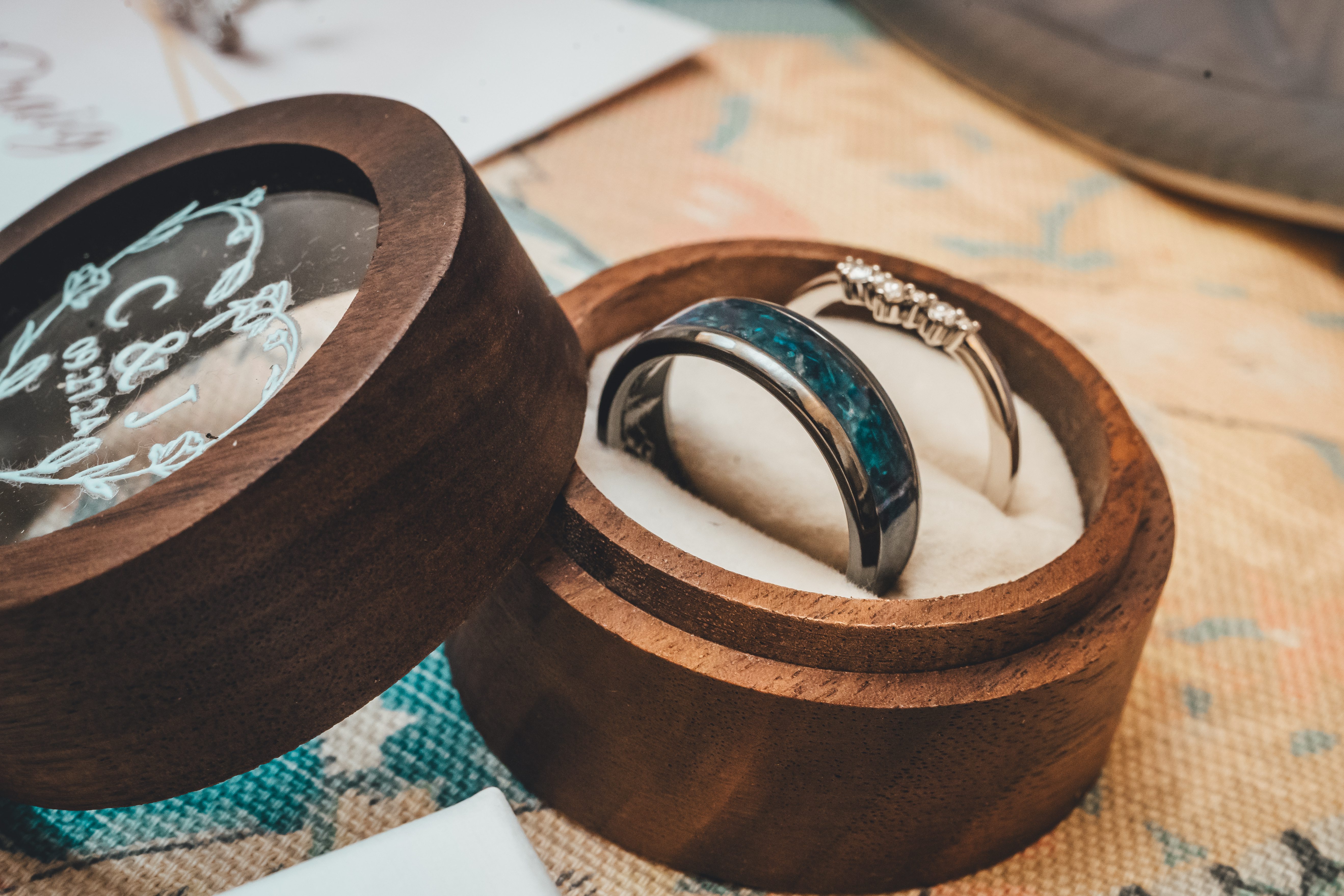 Two wedding rings displayed in wooden ring boxes on a neutral fabric surface. The groom's band features a teal/dark enam