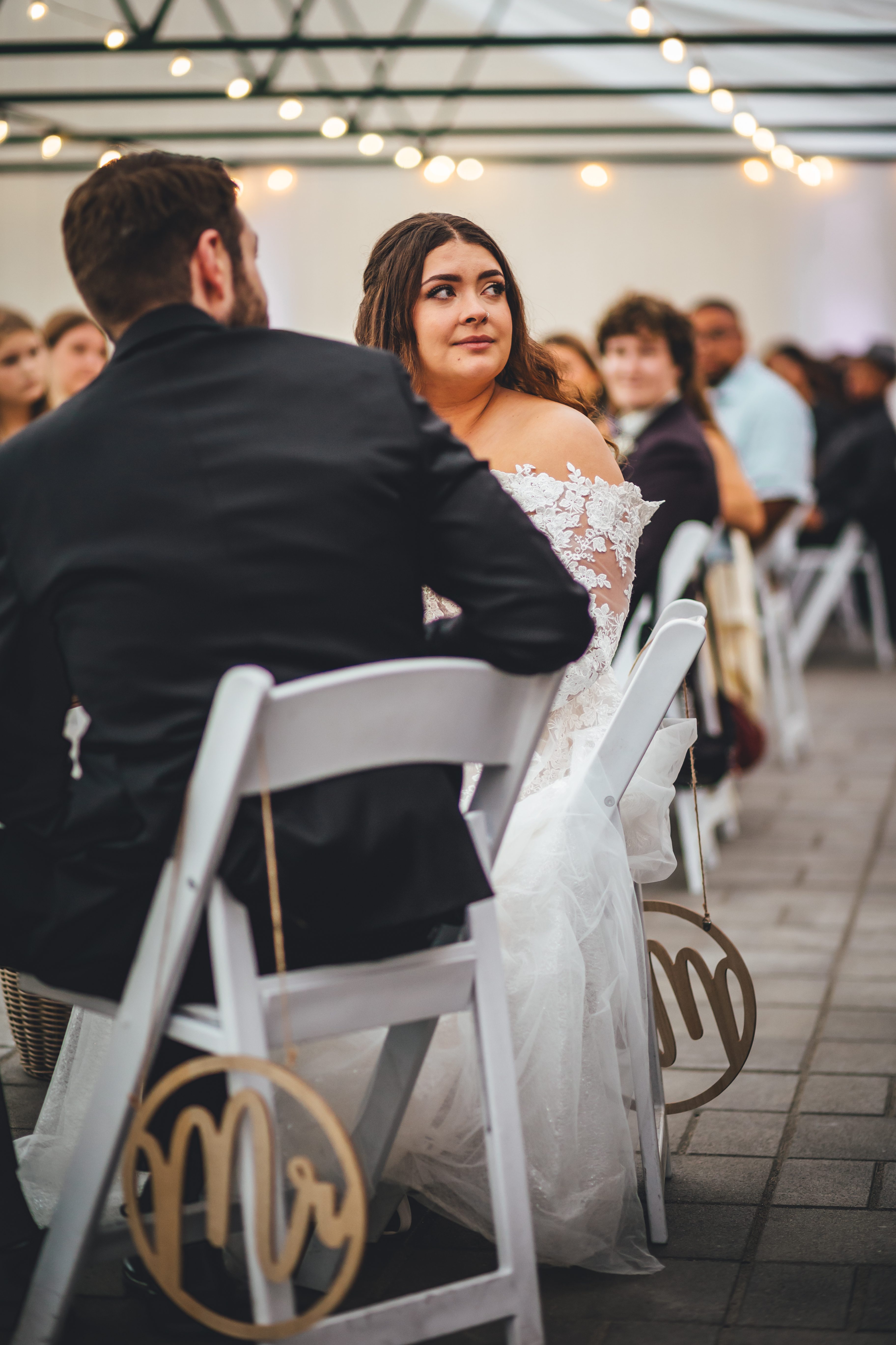 The bride sits in the ceremony space wearing a white lace off-shoulder gown, gazing upward as the groom (in black suit) stands beside her. Guests are seated in 