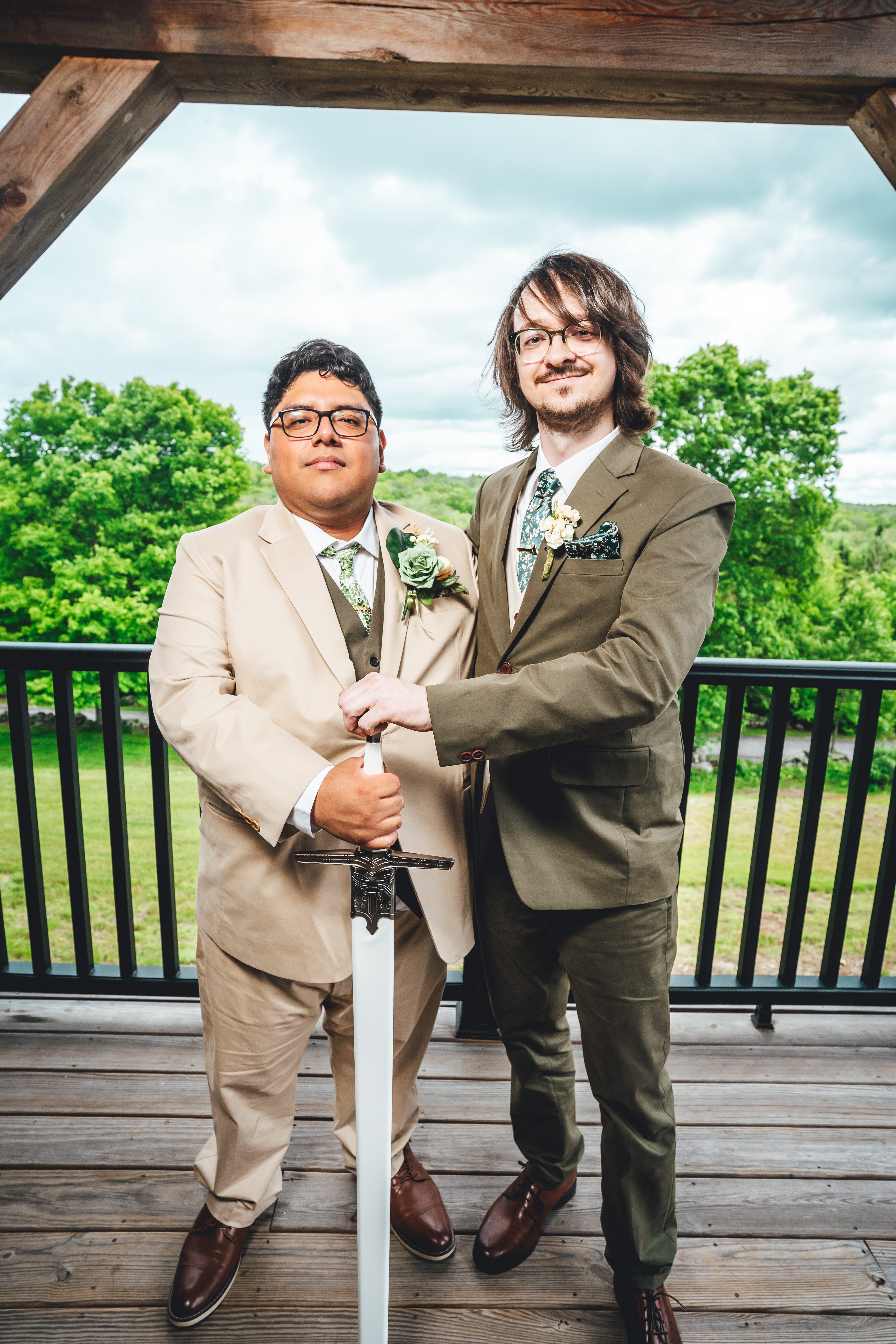 Two grooms in coordinating earth-tone suits pose together on an outdoor wooden deck beneath a rustic timber frame, holdi