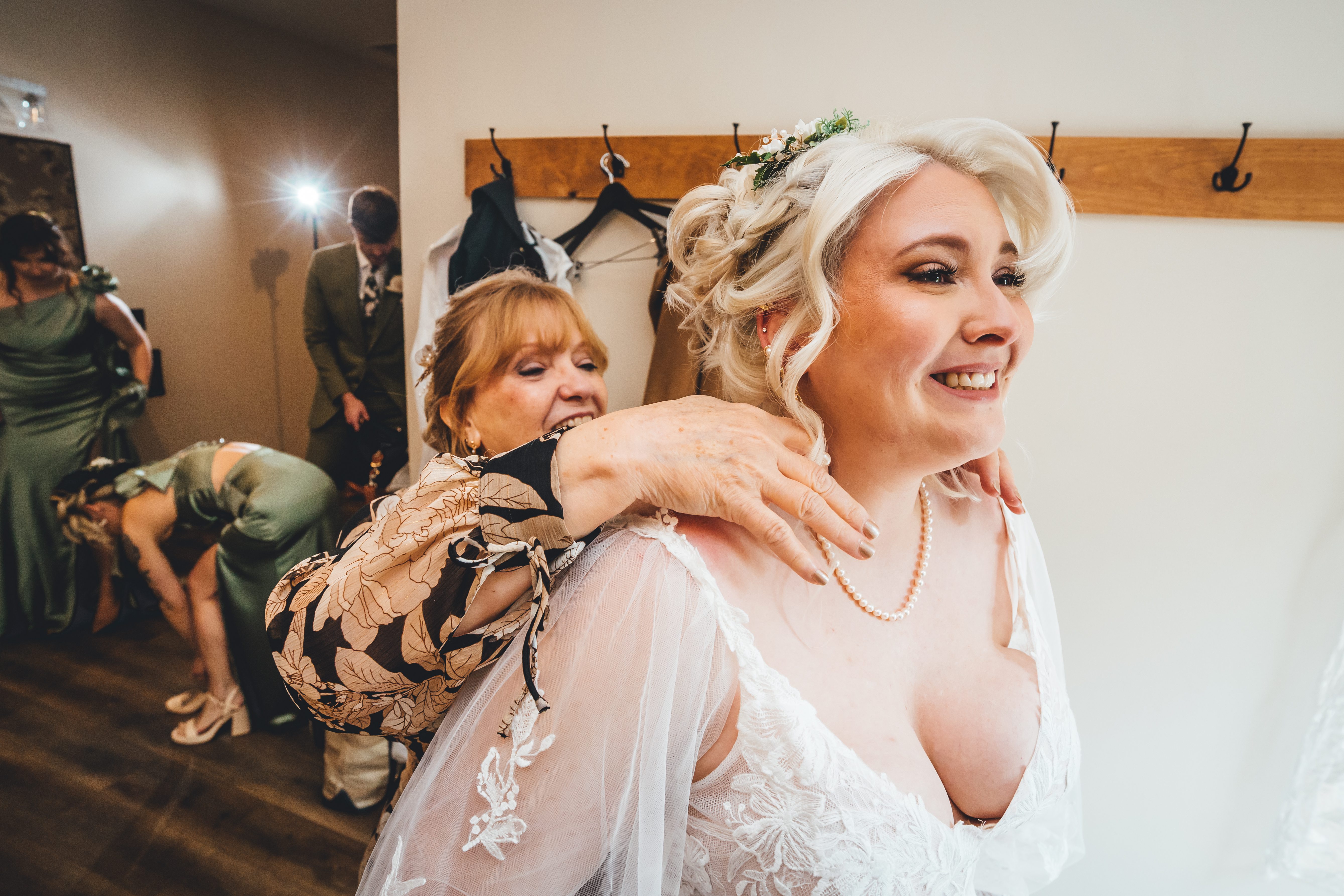 Bride in white lace dress with blonde wavy hair and flower crown smiles radiantly while holding a large animatronic dragon puppet on her shoulder. A woman in ol
