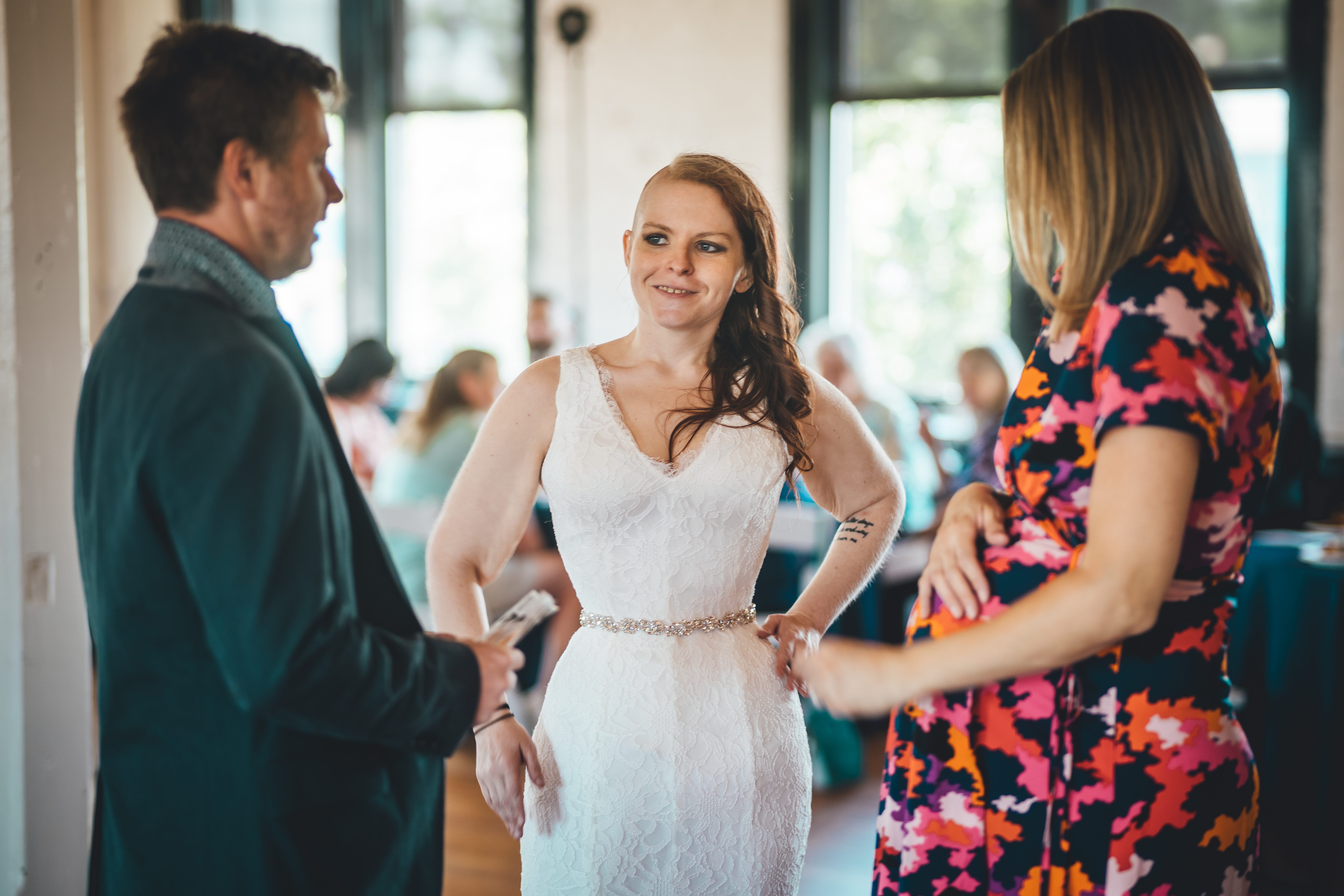 The bride in an elegant white lace gown with beaded waist belt stands smiling while conversing with a man in a dark teal sweater and a woman in a vibrant multic