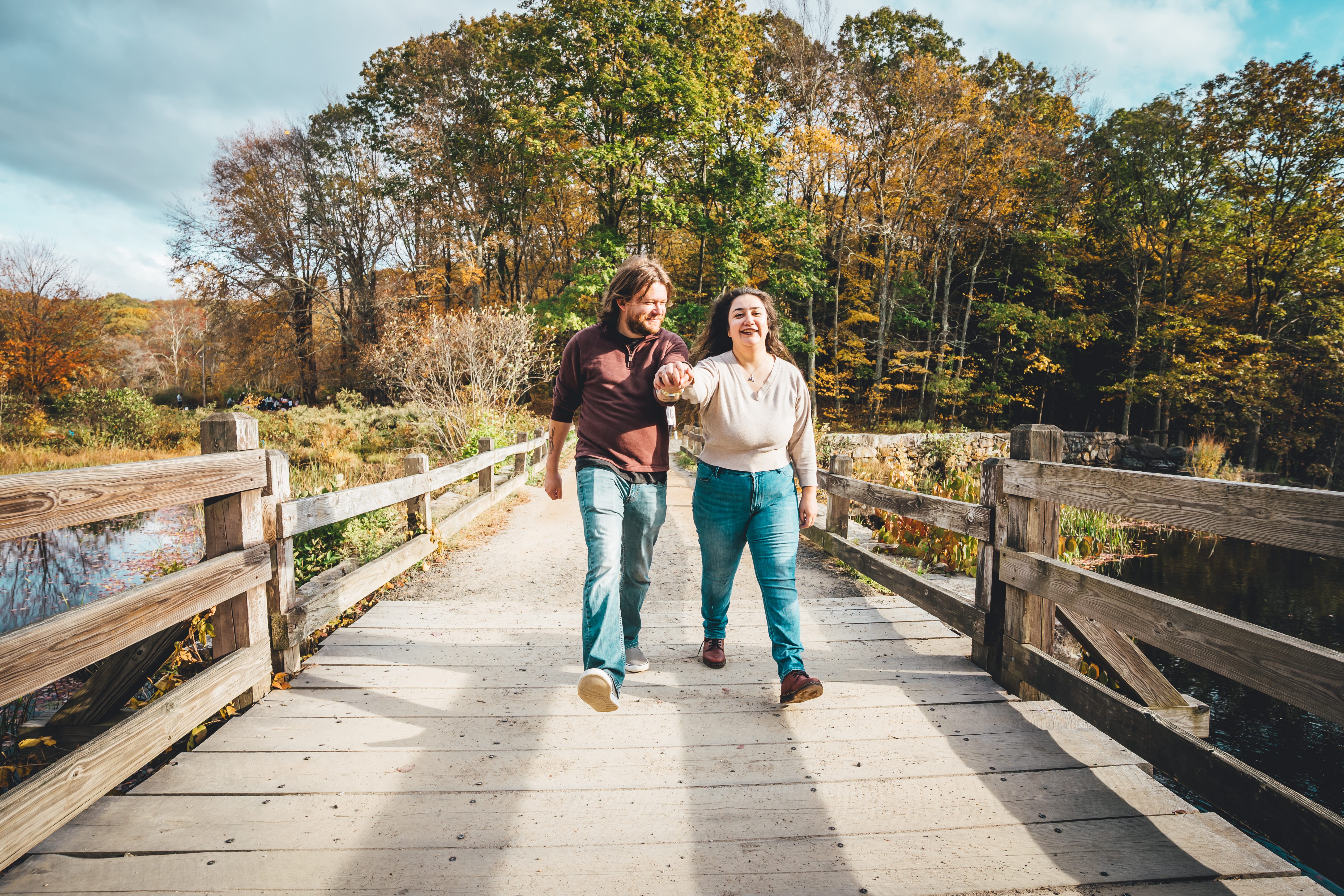 An engaged couple walks hand in hand across a wooden bridge in autumn woods