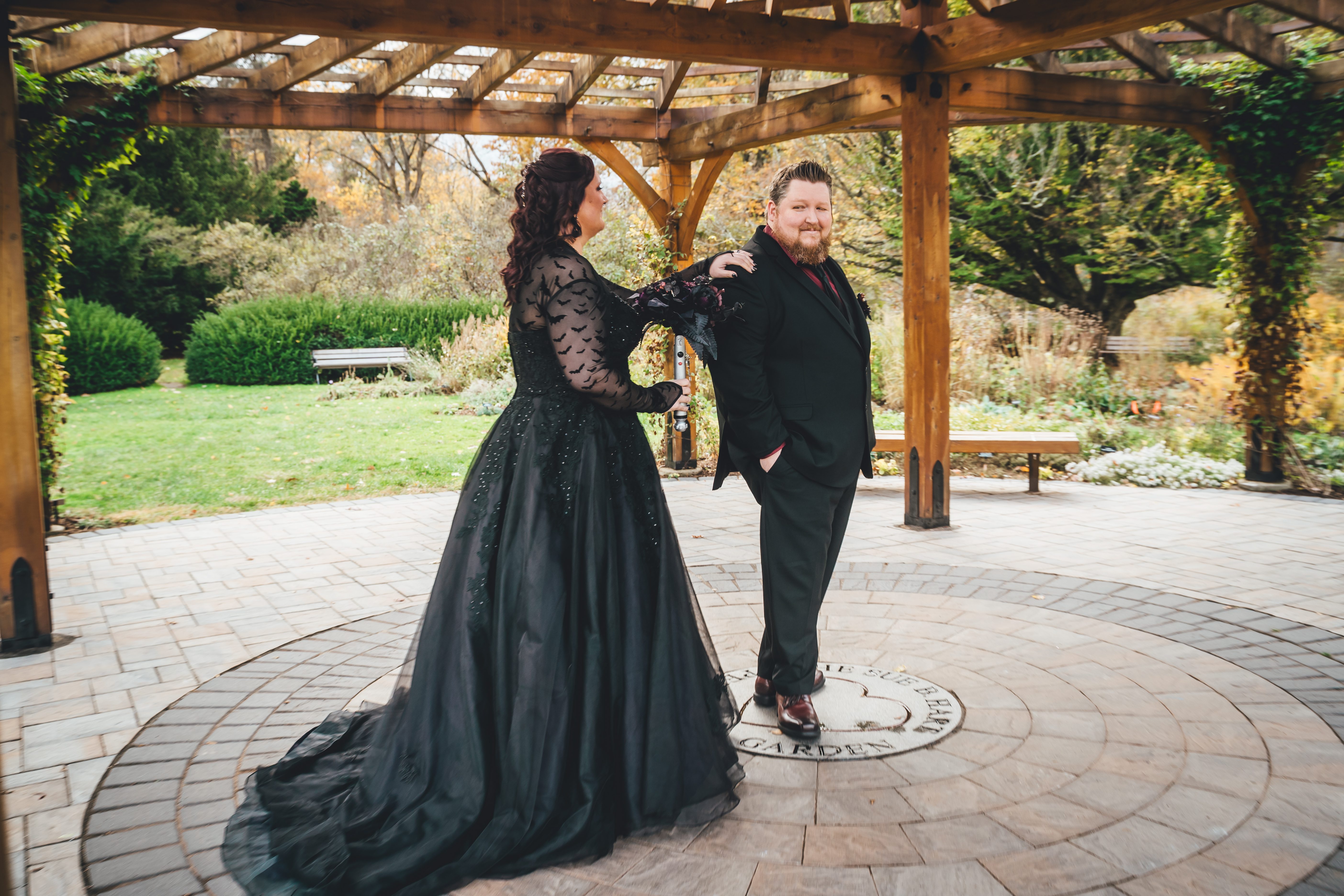 A couple poses together under a wooden pergola on a paver patio. The bride wears an elaborate black gown with sparkly embellishments and scale-patterned lace sl