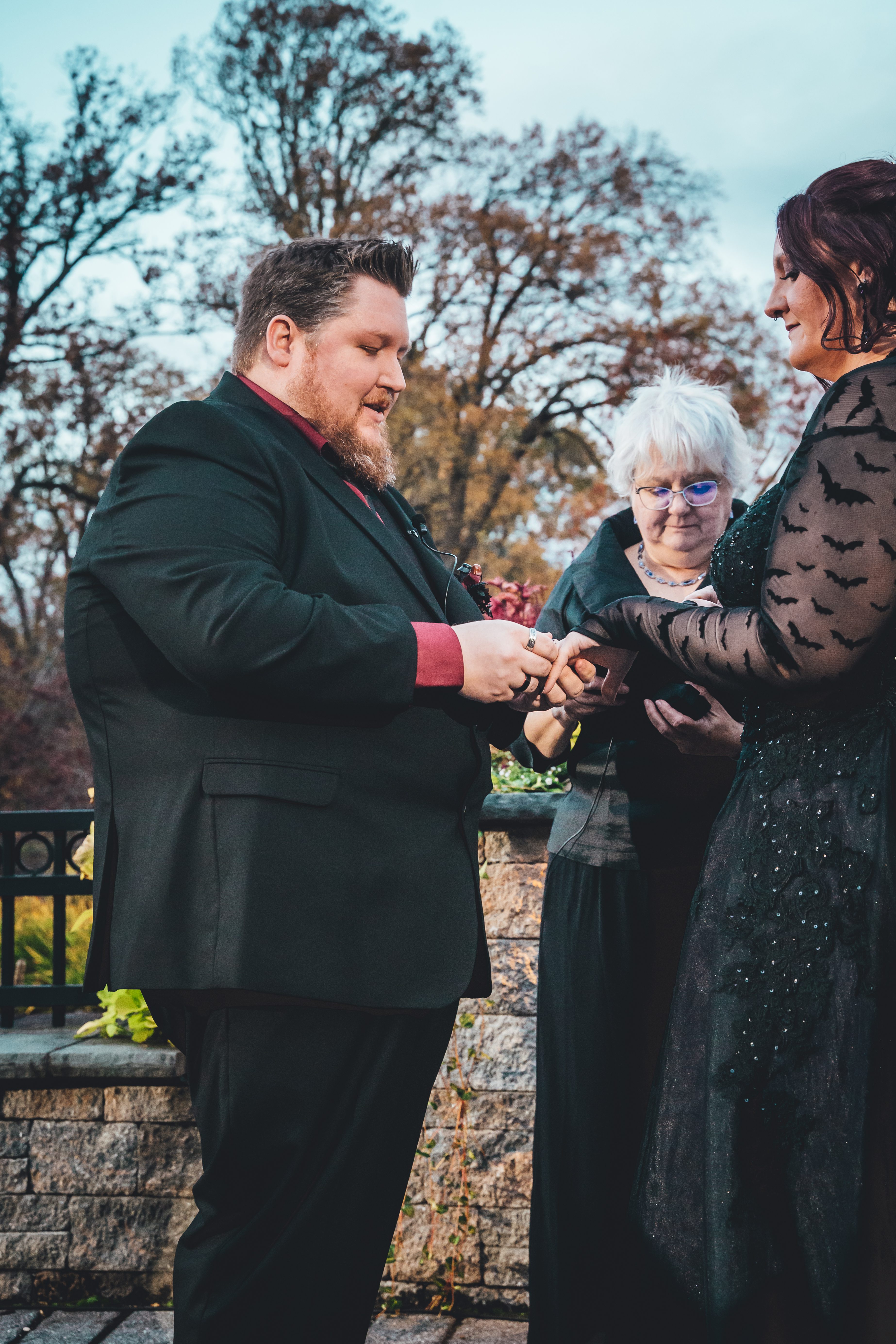 Groom in dark suit with burgundy shirt cuffs exchanges rings with bride during outdoor ceremony; elderly woman in glasses and bat-print dress stands as witness,