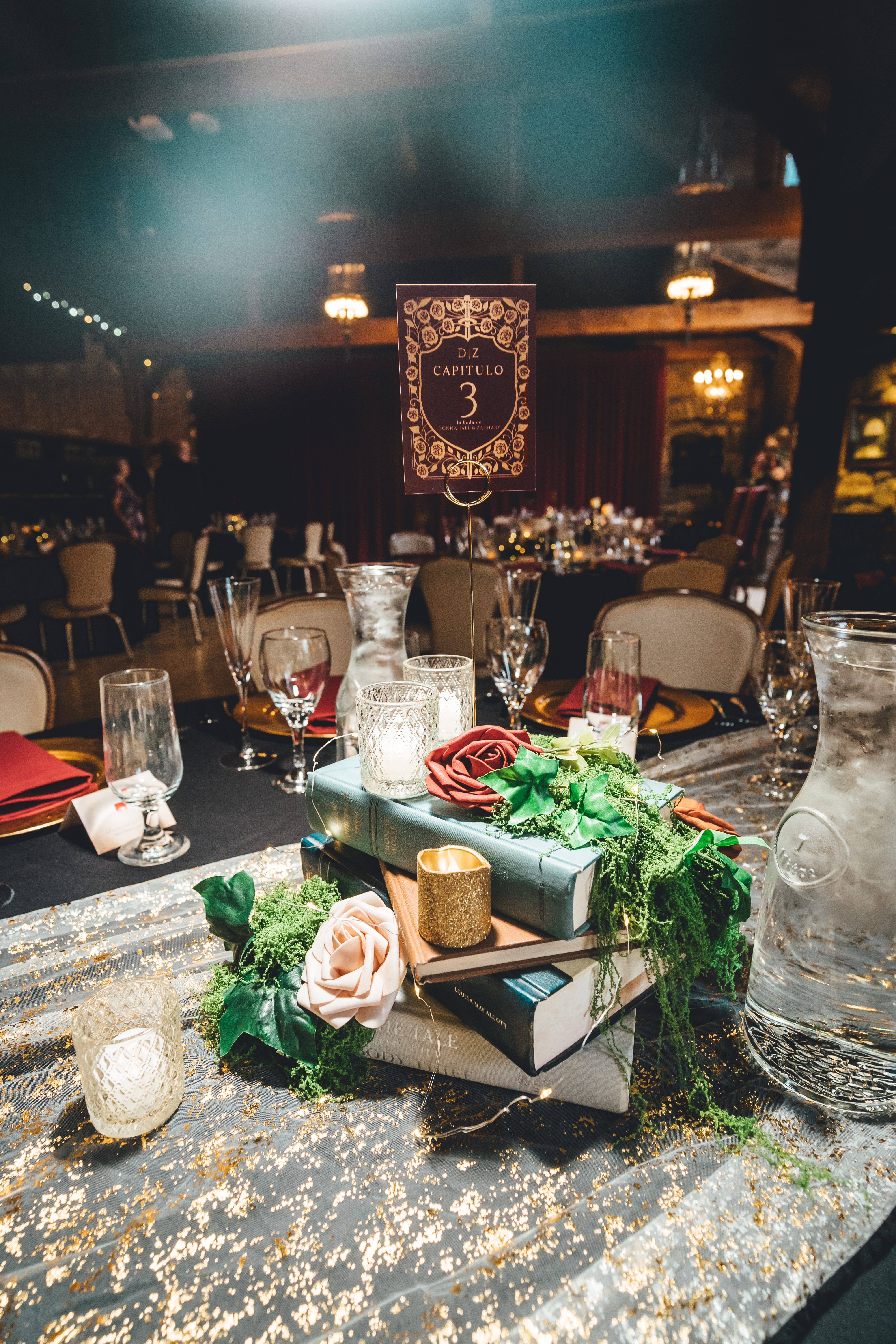 An elegantly styled reception table centerpiece featuring stacked books with blush roses and greenery, ornate table numb
