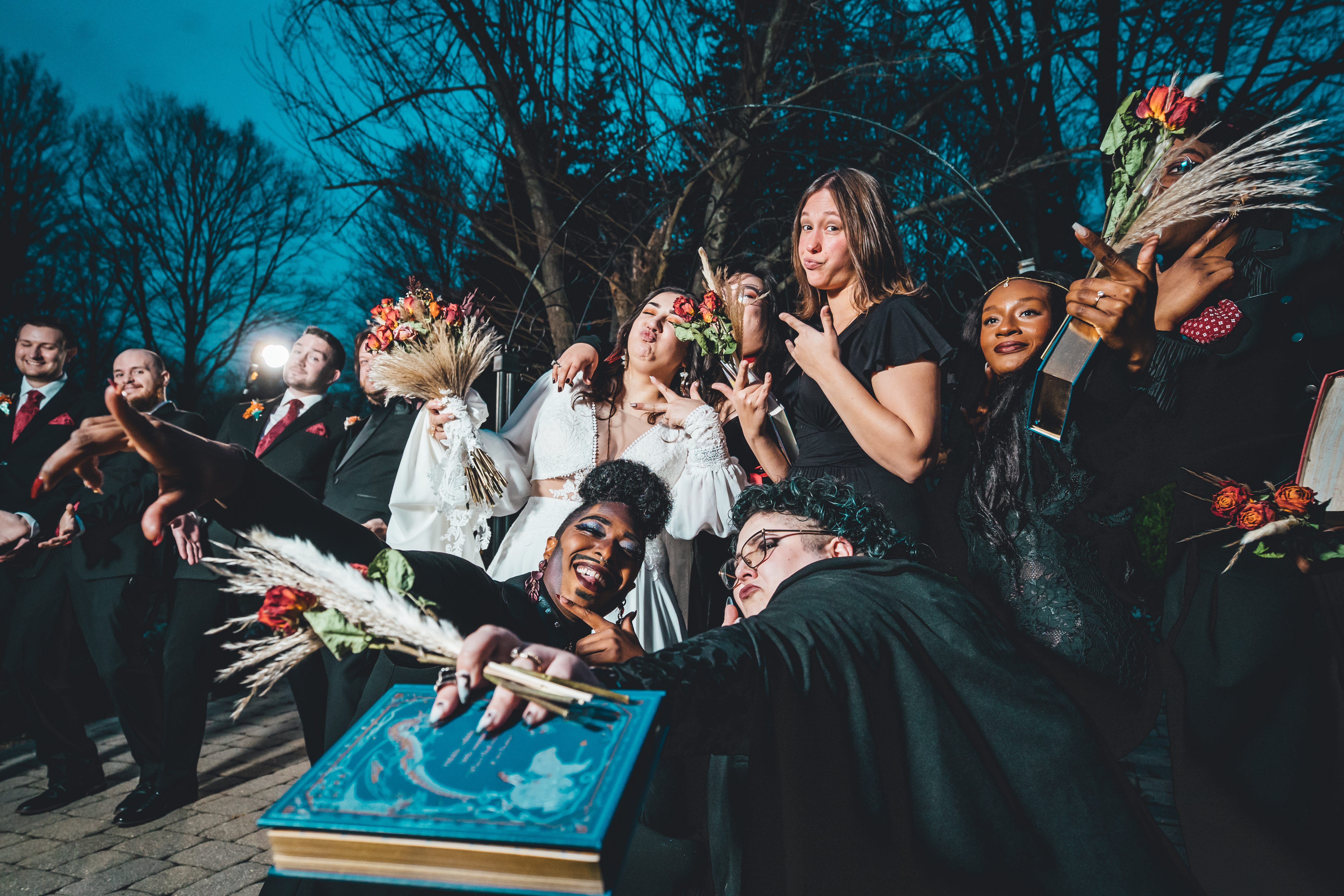 A lively wedding party celebrates at dusk with dramatic flair. The bride in white stands center among bridesmaids in bla