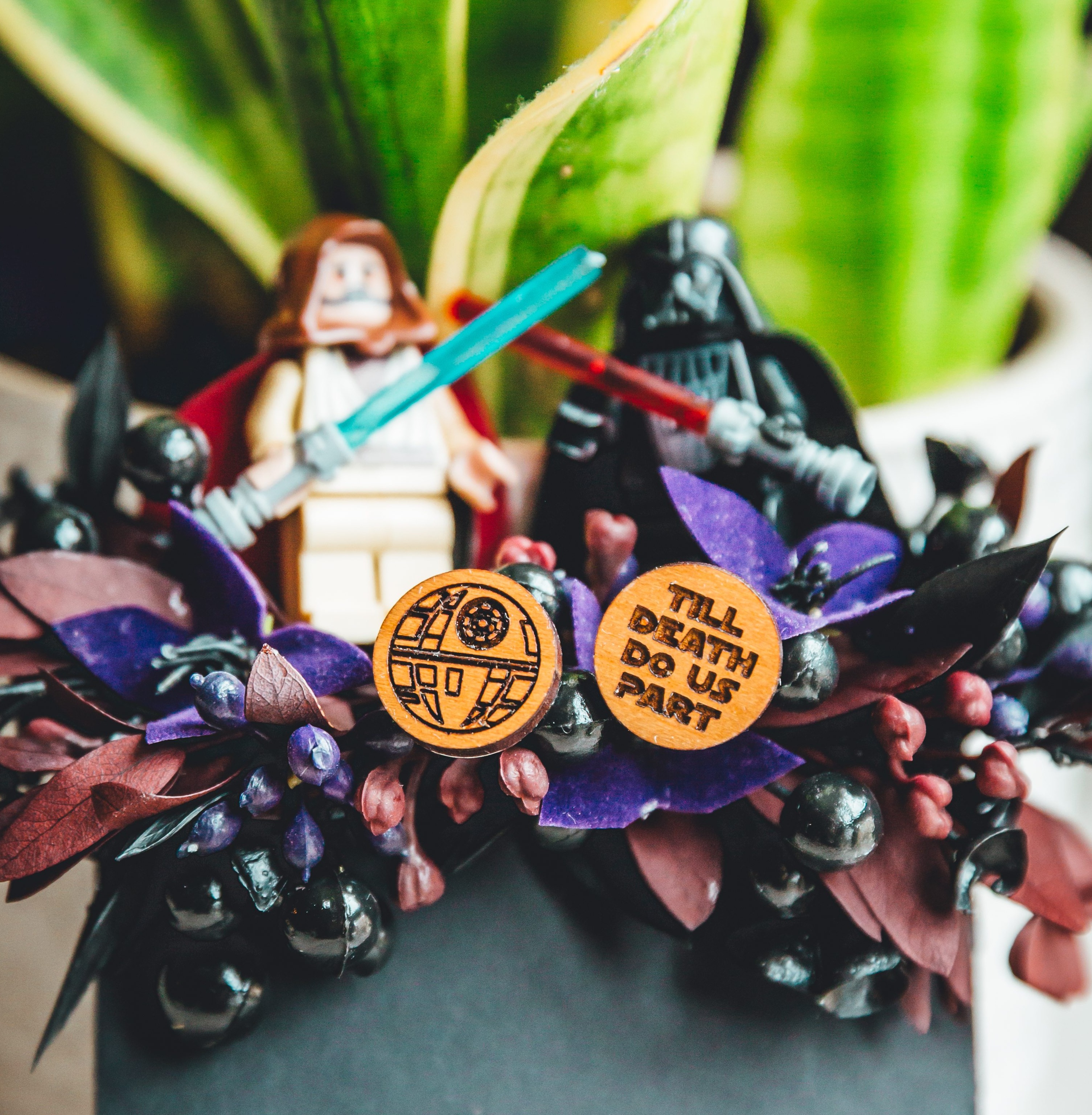 A close-up of a wedding bouquet featuring a teal vase with green foliage, dark purple and burgundy flowers, and Star Wars-themed decorative elements including Darth Vader and Luke Skywalker figurines with lightsabers, plus two custom wooden coins with Death Star and 'I FIND YOUR LACK OF FAITH DISTURBING' text.