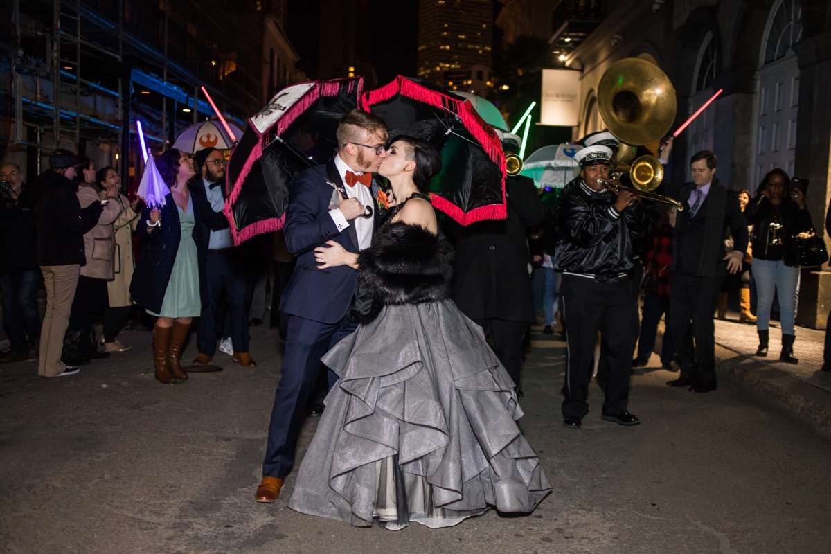 A couple in formal attire kisses while guests raise lightsabers behind them